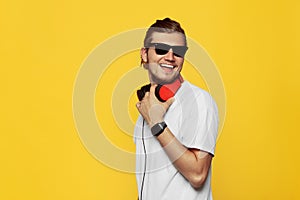 Cheerful hipster guy smiles happily, has excited expression wearing white t shirt, sunglasses and headphones, isolated over yellow