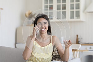 Cheerful Hispanic girl speaking on cellphone, sitting on home couch