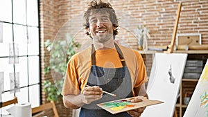 A cheerful hispanic man in a studio, painting on a palette, radiates creativity and joy in a casual setting photo