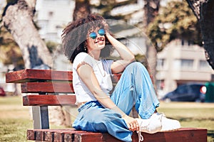 Cheerful hispanic woman wearing sunglasses and sitting on a park bench outside. Carefree young woman with a curly afro