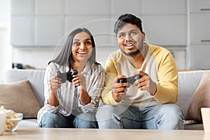 Cheerful Indian Couple At Home Playing Video Games With Joysticks