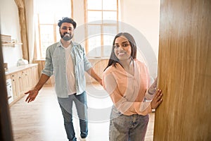 Cheerful indian couple inviting guests to enter home, happy people standing in doorway of modern flat