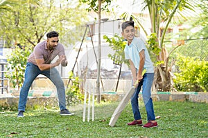 Cheerful indian father encouraging son from behind while playing cricket game at park - concept of Motivation, Guidance