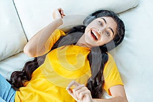 Cheerful Indian girl in wireless headphones dancing on couch to her favorite music playlist.