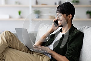 Cheerful indian guy independent contractor with laptop having phone conversation