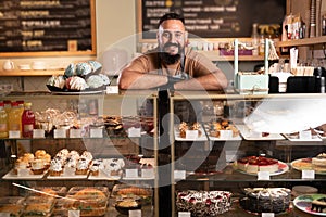 Cheerful indian male baker welcoming you at his bakery store, bearded male baker smiling to the camera proudly leaning