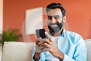 Cheerful Indian Man Using Mobile Phone Surfing Internet Sitting Indoor