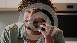 Cheerful Indian young man with curly hair talking phone in home kitchen. Happy friendly Hispanic guy talk with friends