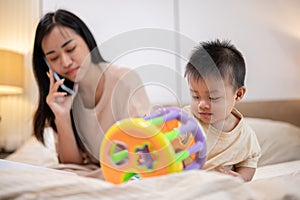 A cheerful, innocent Asian baby boy is playing toys in bed while his mom is talking on the phone