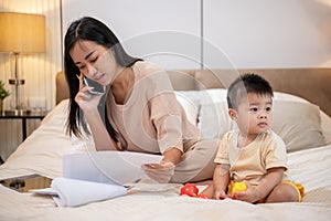 A cheerful, innocent Asian baby boy is playing toys in bed while his mom is talking on the phone