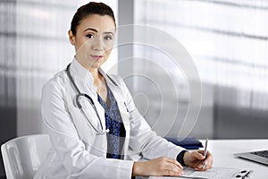 Cheerful intelligent woman-doctor is using a clipboard and writing some medication names, while sitting at the desk in