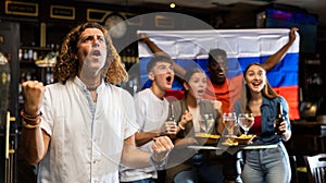 Cheerful international soccer football fans waving Russian flag and drinking beer, eating chips in the sport bar