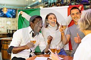 Cheerful international sport fans waving Italian flag and drinking beer in the sport bar