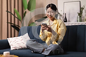 Cheerful Japanese Millennial Woman Using Cellphone Wearing Earphones At Home