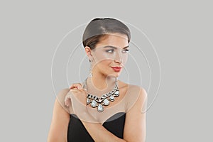 Cheerful jewelry model woman with dark brown hair and shiny fresh skin wearing diamond earring and necklace posing against grey