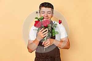 Cheerful joyful male florist wearing brown apron standing isolated over beige background at flowers shop smelling beautiful red