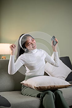 A joyful Asian woman is enjoying the music and dancing while sitting on a sofa in her living room