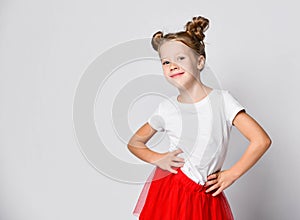 Cheerful kid girl in red fatin skirt and white t-shirt is standing holding hands on her waist to us having fun dancing