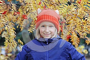 Cheerful kid girl smiling, the child is dressed in a funny knitted warm hat with ears, looks like a fox. Autumn, outdoors portrait