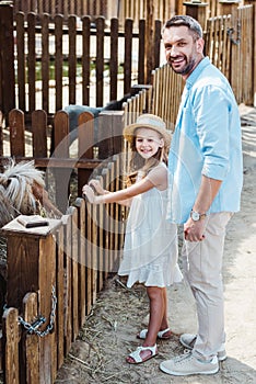 cheerful kid and man smiling while standing near pony in zoo.