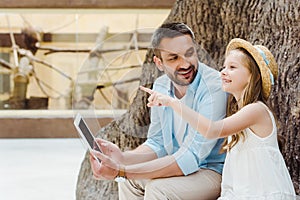 cheerful kid in straw hat pointing with finger near father with digital tablet.