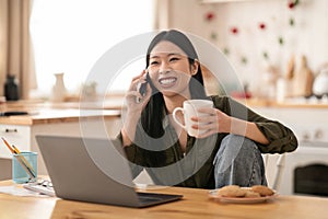 Cheerful korean woman using laptop at kitchen, talking on phone