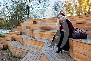 A cheerful lady sits on a summer theater bench, made of wood in a burgundy coat and biret, an adult smiles at the camera