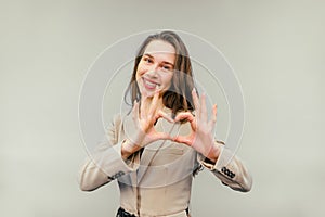 Cheerful lady in smart casual clothes stands on a beige background, looks at the camera with a smile on his face and shows a heart