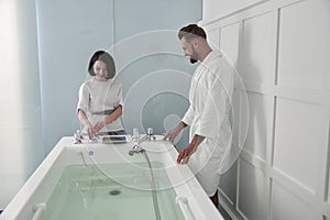 Cheerful lady tunes hydro massage tub full of clear water to mature man in spa salon