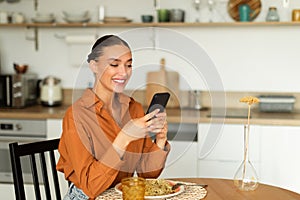 Cheerful lady using smartphone while sitting at kitchen table and having lunch, texting on mobile phone, free space