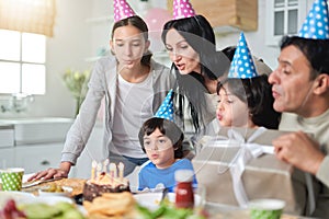 Cheerful latin american family wearing birthday caps, blowing candles on a cake while celebrating birthday together at