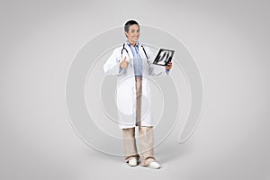 Cheerful latin doctor woman wearing stethoscope holding chest xray and doing ok sign, grey background, full length shot