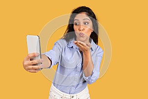 Cheerful latin millennial brunette lady in casual make air kiss at smartphone, make selfie