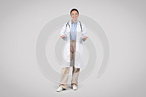 Cheerful latin woman doctor smiling to camera posing, wearing white uniform, grey background, full length