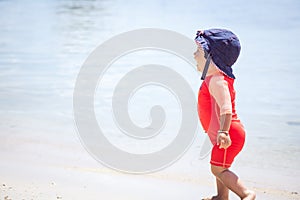 Cheerful laughing baby in the bright sun protective suit running on beach with sea on background