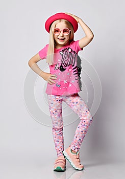 Cheerful laughing blonde kid girl in round sunglasses, t-shirt with zebra print stands holding hand on her pink hat