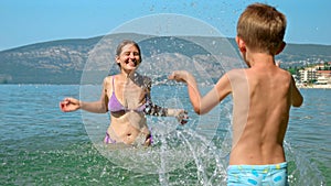 Cheerful laughing mother splashing sea water on her little son relaxing on beach