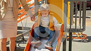 Cheerful laughing toddler boy holding mother's hand and sliding down on the playground
