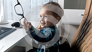 Portrait of cheerful laughing toddler boy sitting in office and playing with eyeglasses