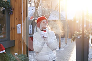 Cheerful laughing woman walks through the city`s decorated streets at Christmas time with mug of hot cocoa.