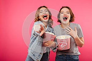 Cheerful laughing women friends eating popcorn watch film pointing.