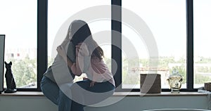 Cheerful lesbian couple hugging on windowsill and smiling. Side view portrait of joyful young Caucasian women enjoying