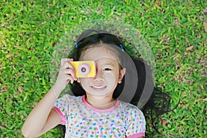 Cheerful little Asian girl takes photo with colorful digital camera lying on green lawn background