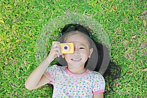 Cheerful little Asian girl takes photo with colorful digital camera lying on green lawn background