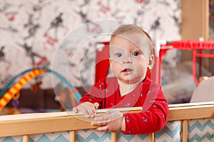 A cheerful little baby up to a year old stands in his crib in the children`s playpen and looks at the camera in surprise, the emo