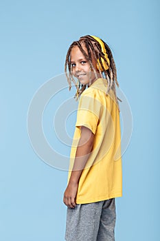 Cheerful little boy with african dreads listening music in headphones has pleasant smile over blue background.