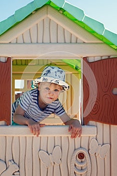 A cheerful little boy in a hat, inside a toy little house, he looks out the window with the shutters straight into the camera