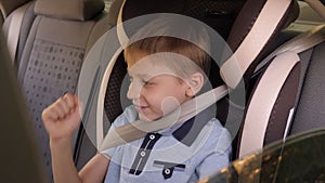 A cheerful little boy is sitting in a child car seat in the car and dancing.