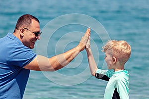 Cheerful little boy in surfing swimwear giving high five to dad on beach expressing trust and confidence