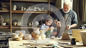 Cheerful little boy is throwing pieces of clay on work table while helping his grandfather in potter`s workshop. Happy